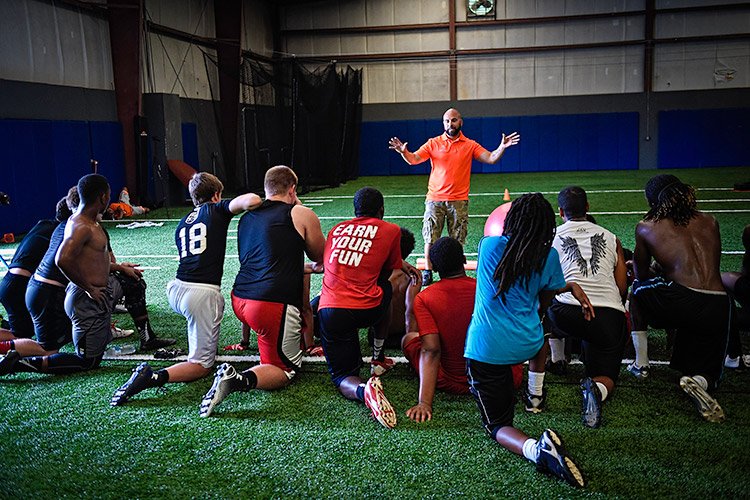 Wounded warrior Carlos De León, dressed in an orange Wounded Warrior Project polo and cameo cargo shorts, delivers a presentation to fellow warriors and family members in an indoor football gym.