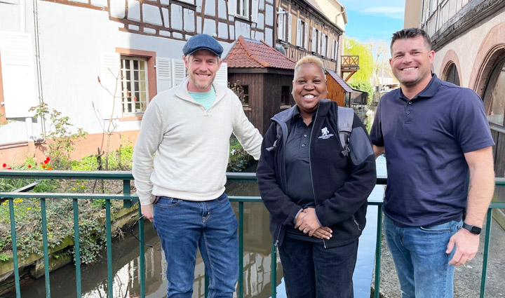 Wounded Warriors John Rego, Donna Pratt, and Mike Larson stand in front of a river in between two buildings while smiling.